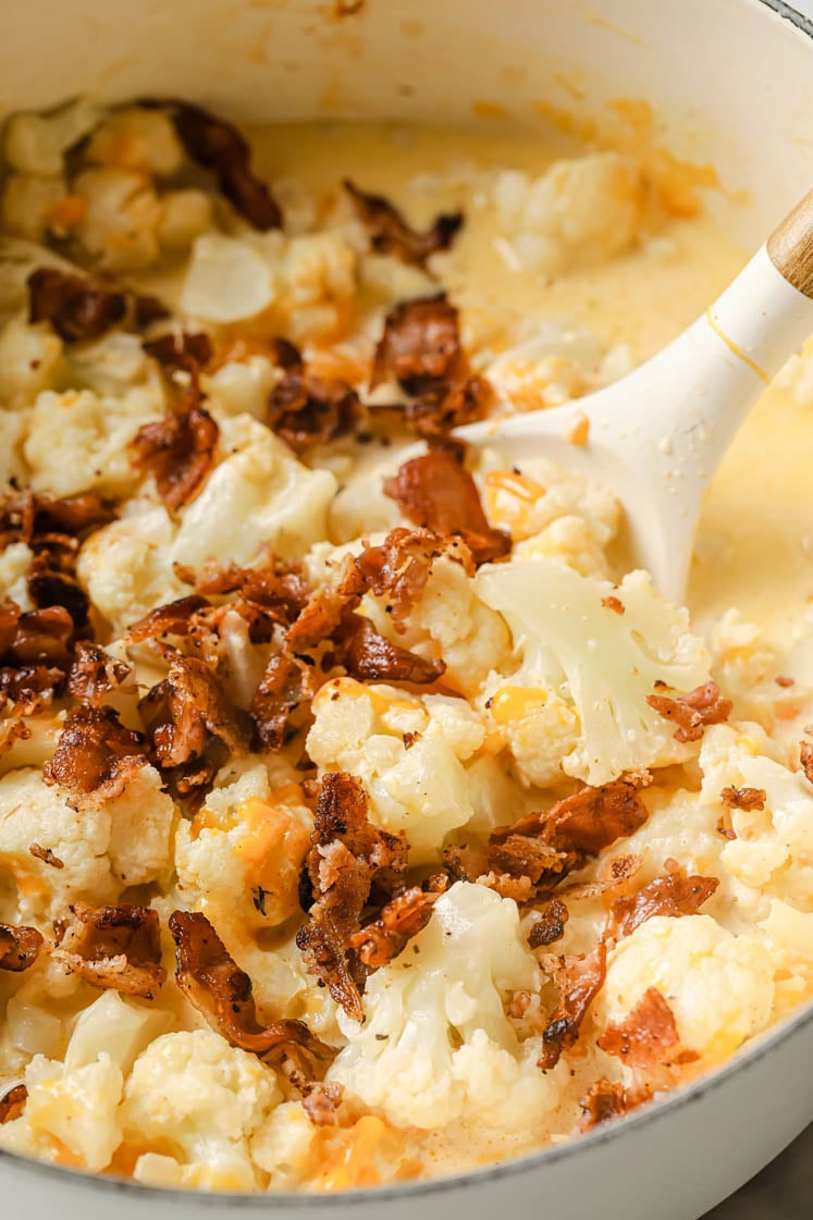 Overhead shot of a large pot with mixed cauliflower florets, chicken broth, spices, heavy cream, butter, shredded cheddar and ¾ of the crumbled bacon. Inside the pot, there is a ladle for stirring.