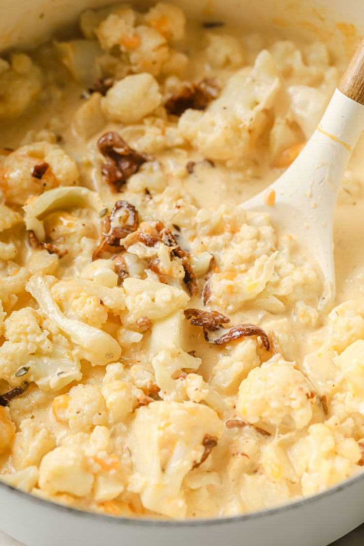 Overhead shot of a large pot simmering with mixed cauliflower florets, chicken broth, spices, heavy cream, butter, melted shredded cheddar and ¾ of the crumbled bacon. Inside the pot, there is a ladle for stirring.