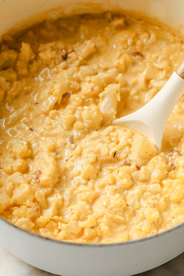 Overhead shot of a large pot simmering with mashed cauliflower florets, chicken broth, spices, heavy cream, butter, melted shredded cheddar and ¾ of the crumbled bacon. Inside the pot, there is a ladle for stirring.