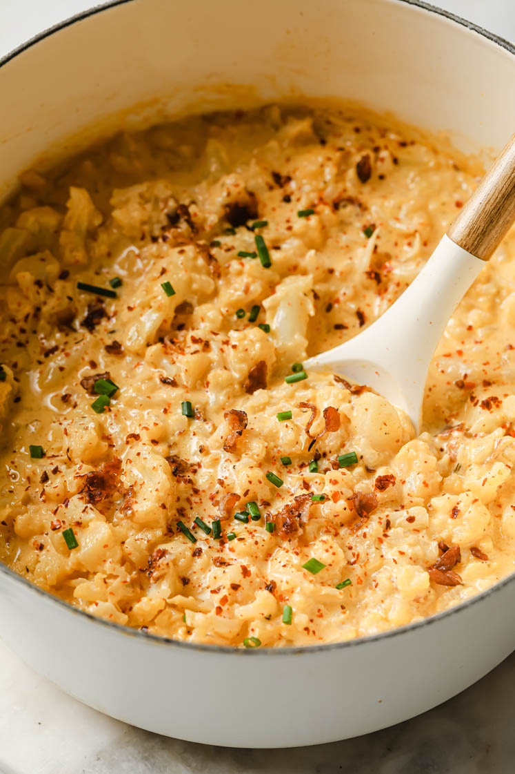 Overhead shot of a large pot with the cauliflower cheddar cheese bacon soup garnished with remaining bacon crumbles, chives and red pepper flakes. Inside the pot, there is a ladle for serving. The pot sits atop a marble countertop.