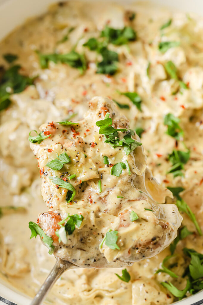 Close-up, overhead shot of a serving spoon with a portion of the Creamy Artichoke Chicken Thighs dish. In the background, we can see a pan with the Creamy Artichoke Chicken Thighs garnished with fresh parsley, freshly cracked pepper, and red pepper flakes.