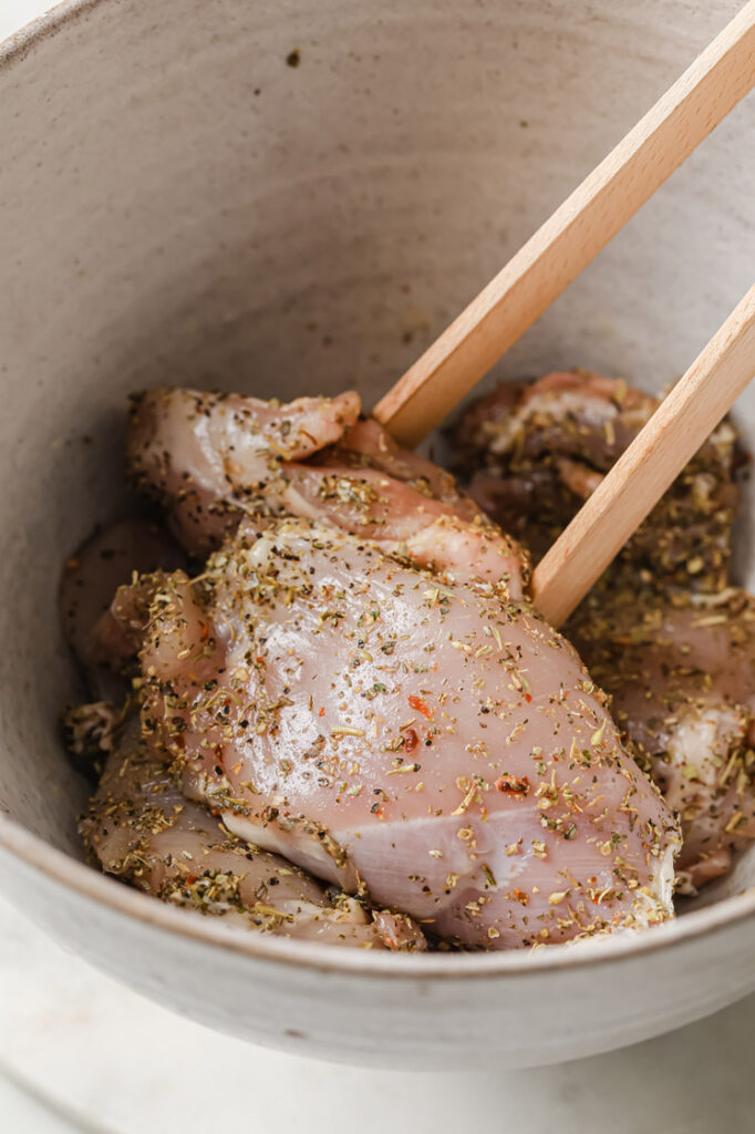 Overhead shot of a large mixing bowl with chicken thighs coated with seasonings using wooden tongs.