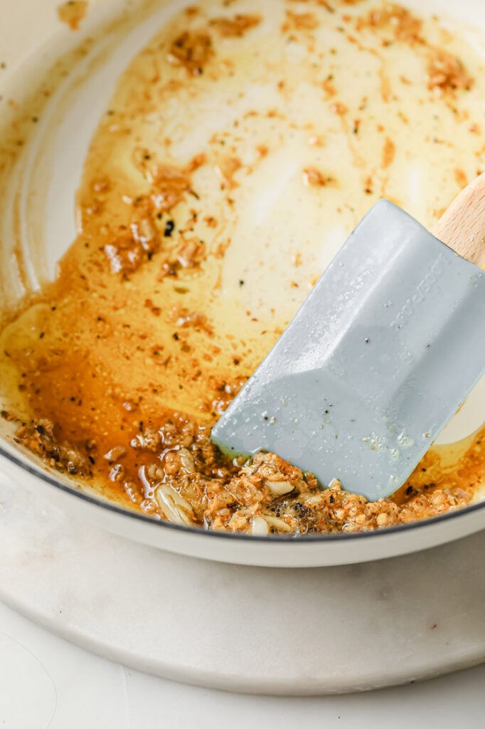 Overhead shot of a large pan with melted butter, browned bits, and minced garlic cooked using a silicone spatula. The pan sits on a white marble trivet. The trivet rests atop a marble countertop.