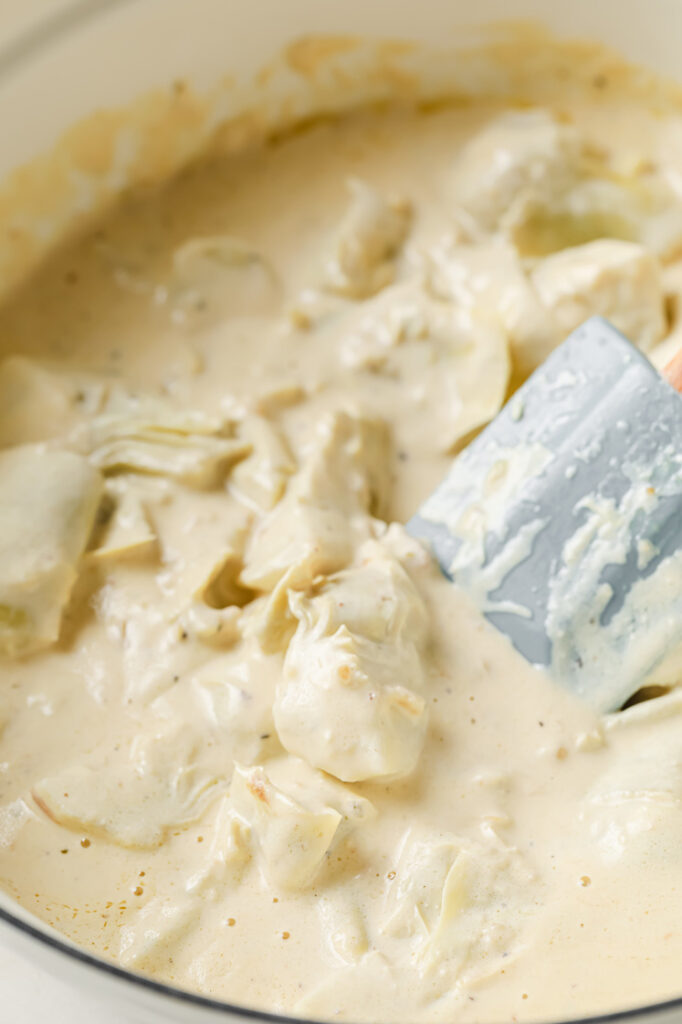 Overhead shot of a large pan with the thickened sauce using melted butter, browned bits, minced garlic, heavy cream, chicken stock, grated Parmesan, salt, and pepper. The artichokes and lemon juice were added and mixed with the sauce using a silicone spatula.