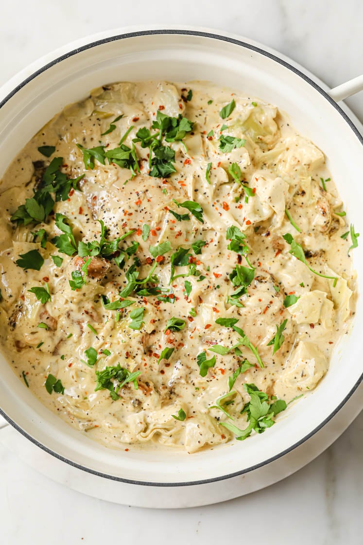 Overhead shot of a large pan with Creamy Artichoke Chicken Thighs, garnished with fresh parsley, freshly cracked pepper, and red pepper flakes. The pan sits on a white marble trivet. The trivet rests atop a marble countertop.