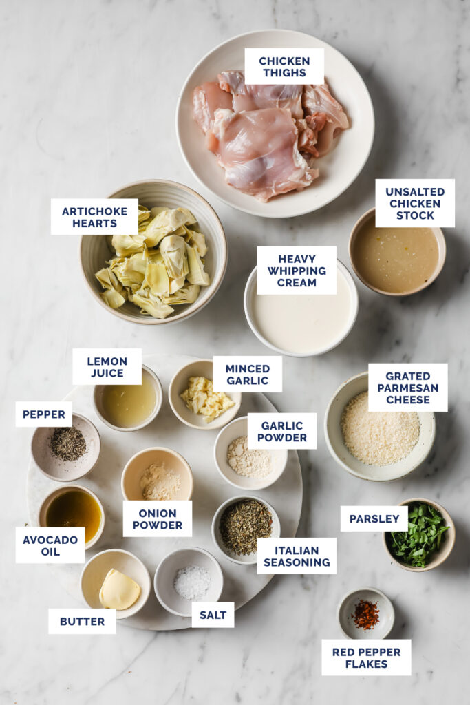 Overhead shot of labeled ingredients for the One-Pan Low-Carb Creamy Artichoke Chicken Thighs, in bowls and plates, atop a marble countertop.