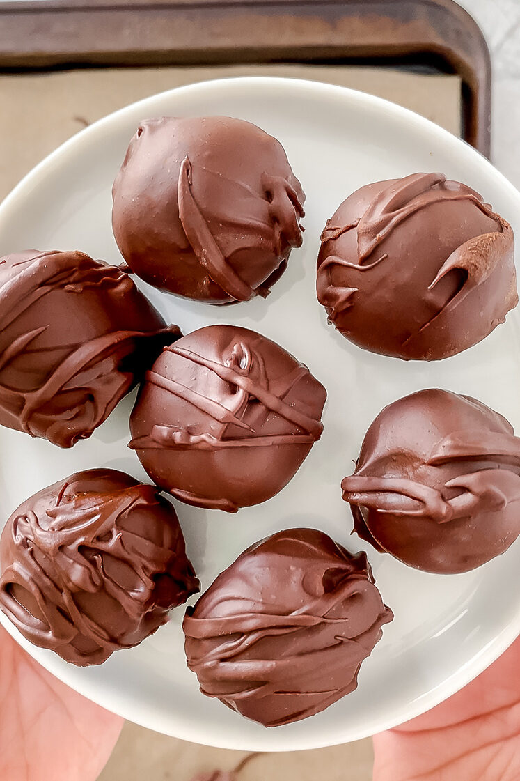 overhead shot of chocolate-covered sunflower seed butter balls on white plate being held in hands above baking sheet atop marble countertop