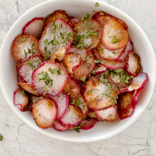 overhead shot of radish chips in white ceramic bowl garnished with dried parsley atop a marble countertop