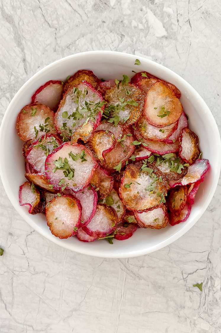 overhead shot of radish chips in white ceramic bowl garnished with dried parsley atop a marble countertop
