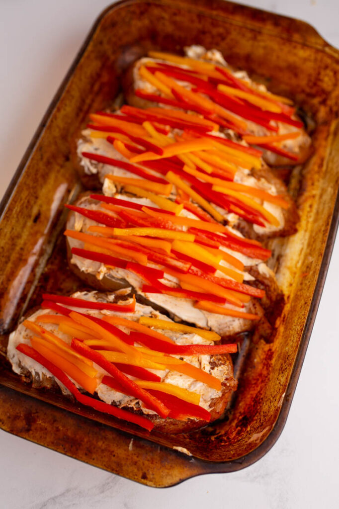 Overhead shot of a baking pan with chicken breasts topped generously with cream cheese and thinly sliced sweet peppers. The baking pan rests atop a marble countertop.
