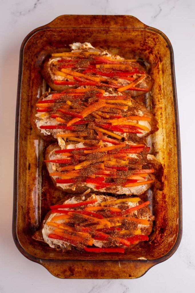 Overhead shot of a baking pan with chicken breasts topped generously with cream cheese and thinly sliced sweet peppers and sprinkled with the remaining spices. The baking pan rests atop a marble countertop.
