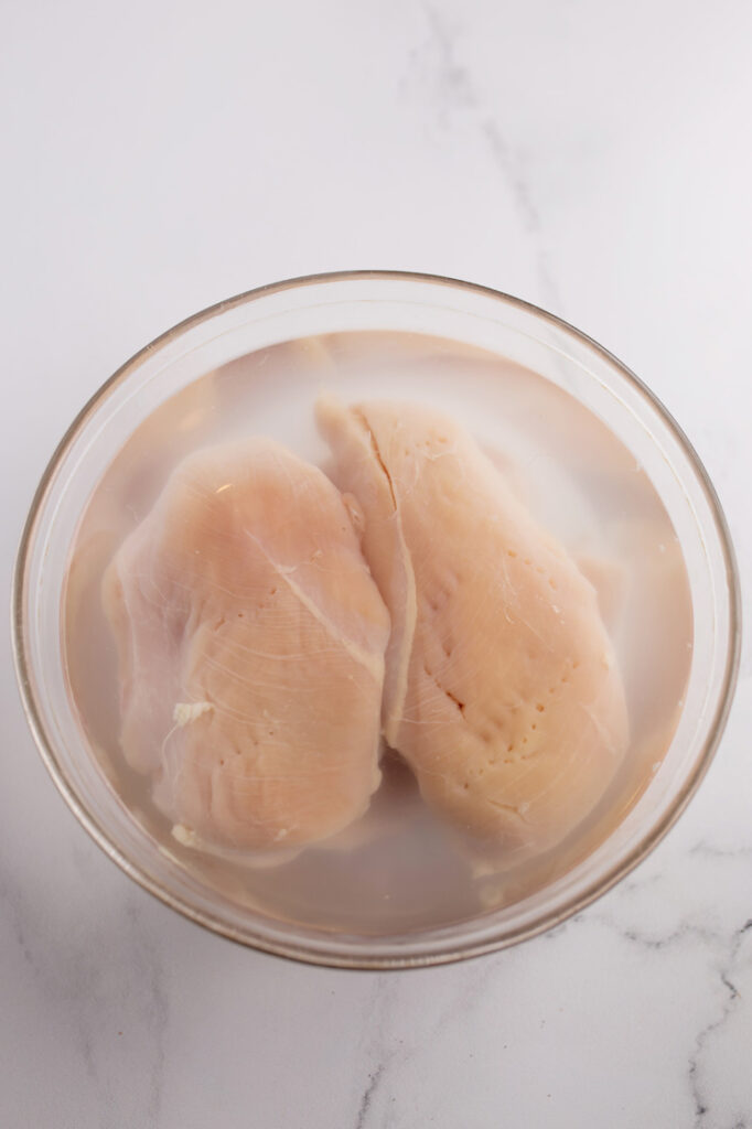 Overhead shot of a glass bowl with brine (made with water and salt) and chicken breasts atop a marble countertop.