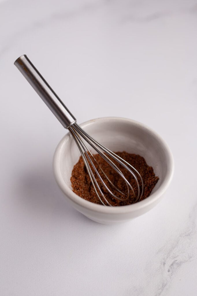 Overhead shot of a small mixing bowl with spices combined by a whisk. The small bowl rests atop a marble countertop.