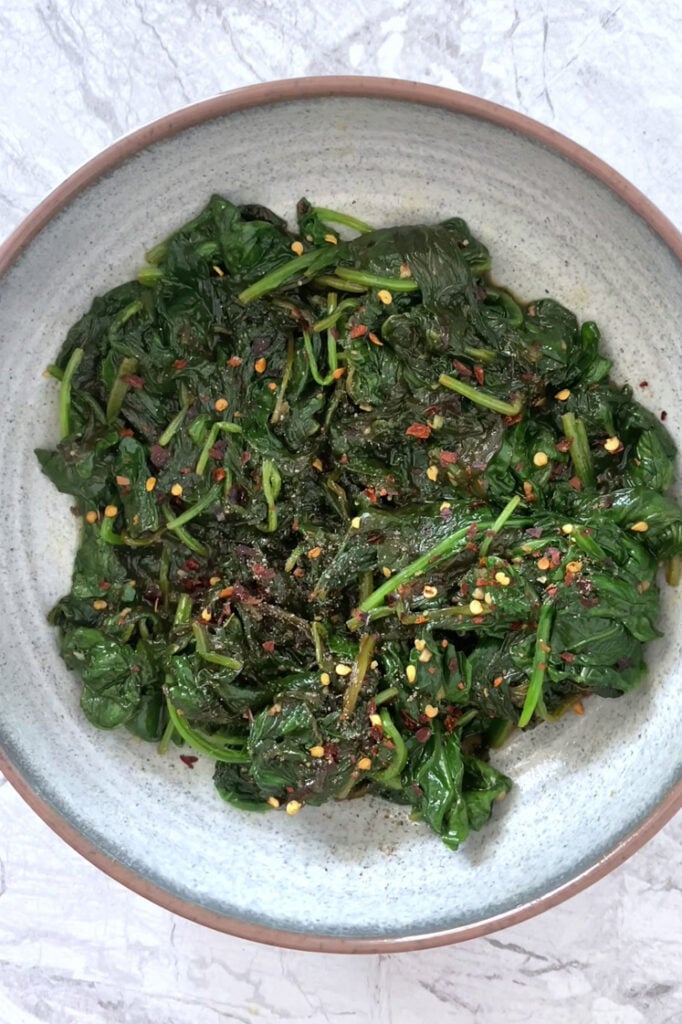 Overhead shot of a serving bowl with the Lemon Garlic Spinach garnished with red pepper flakes and freshly-cracked black pepper atop a marble countertop.
