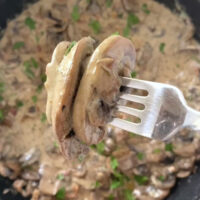 Overhead shot of a fork with a portion of the Creamy Mushrooms and Bacon Skillet over a large skillet with the same dish garnished with fresh parsley and freshly cracked pepper.