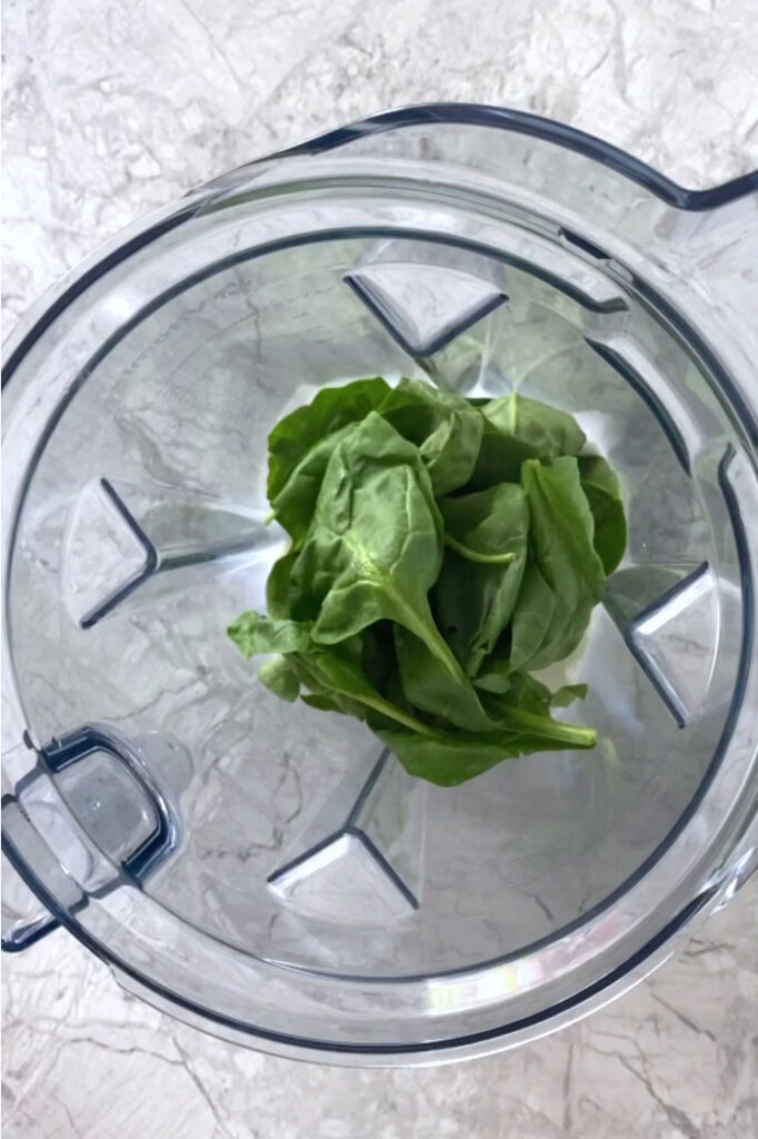 Overhead shot of a blender with coconut milk and fresh spinach atop a marble countertop.
