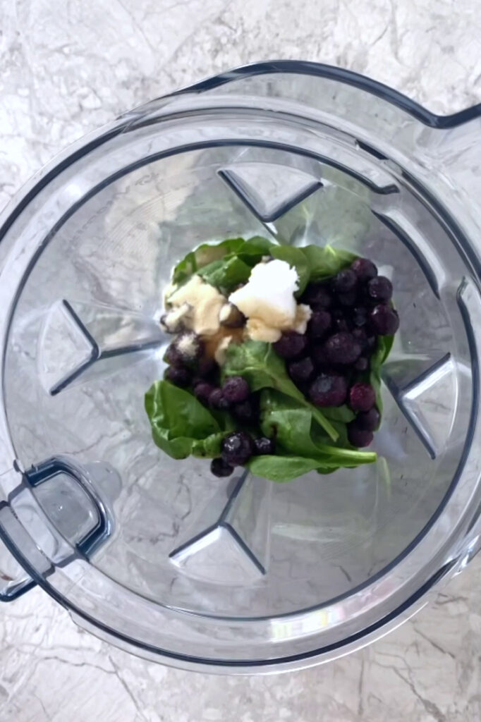 Overhead shot of a blender with coconut milk, fresh spinach, frozen blueberries, coconut oil, gelatin powder, powdered monk fruit sweetener, and vanilla extract atop a marble countertop.