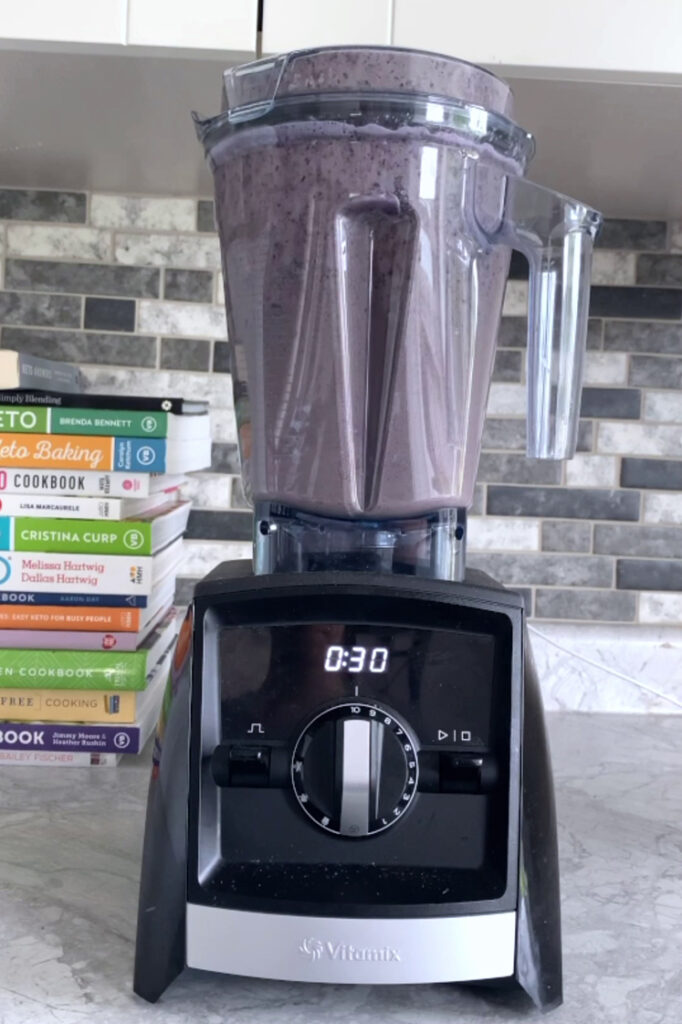 Side shot of a blender with the blended ingredients for the low-carb blueberry spinach smoothie atop a marble countertop.