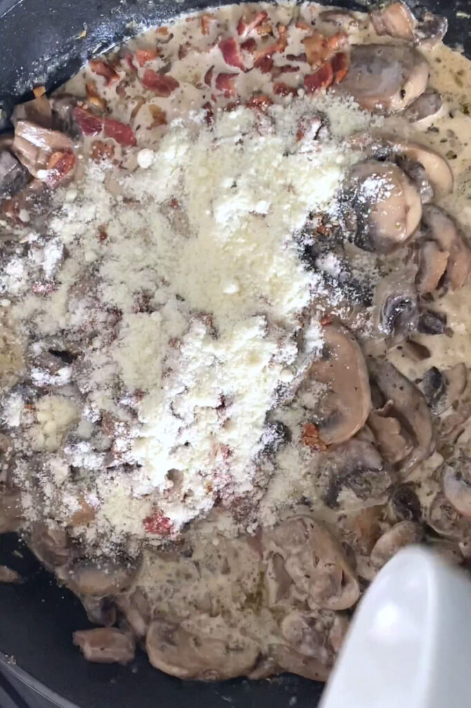 Overhead shot of a large skillet with well-combined bacon grease, melted butter, mushrooms, chicken stock, Italian seasoning, garlic powder, dried thyme, onion powder, heavy cream, salt, and pepper topped with crumbled cooked bacon and grated Parmesan.