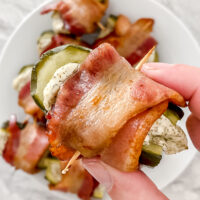 overhead shot of hand holding prepared bacon-wrapped pickle bite atop plates of bites on a marble countertop