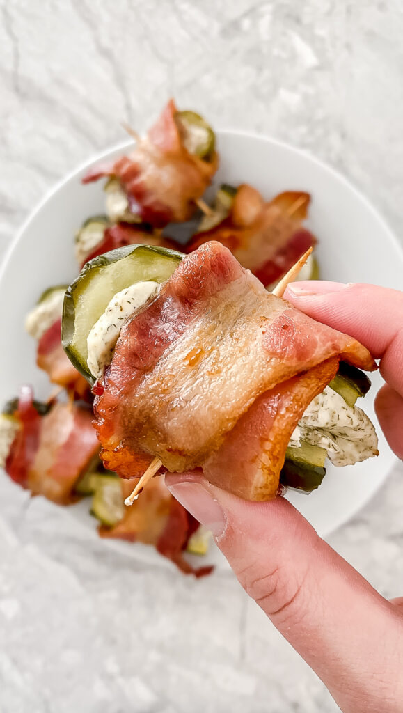 overhead shot of hand holding prepared bacon-wrapped pickle bite atop plates of bites on a marble countertop