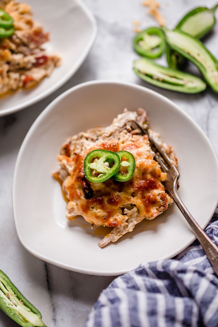 Overhead shot of a serving of Jalapeño Popper Chicken Casserole topped with thinly sliced jalapeños and a fork on a small plate. In the background, there are a few pieces of sliced jalapeños, a blue-striped kitchen towel, and another plate with a portion of the same dish. Everything rests atop a marble countertop.