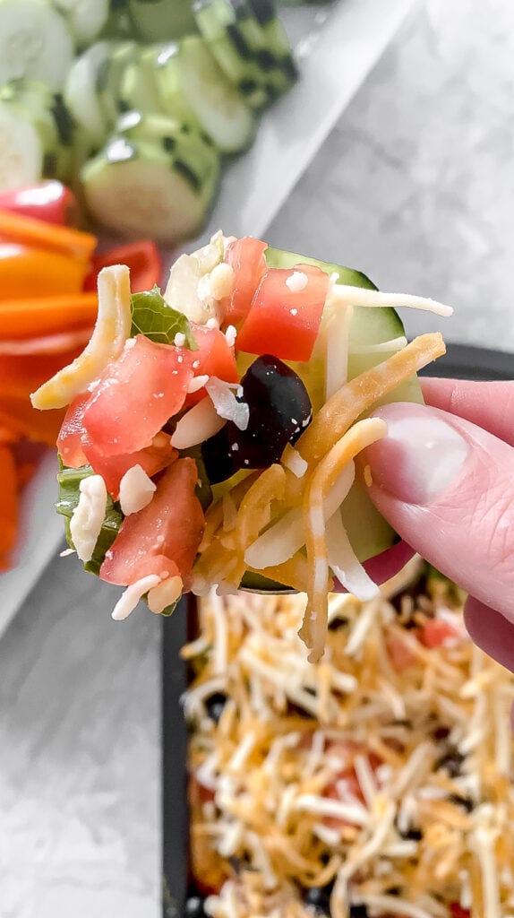 overhead shot of hand holding sliced cucumber topped with low-carb taco dip with pan of dip and dish of vegetables in background