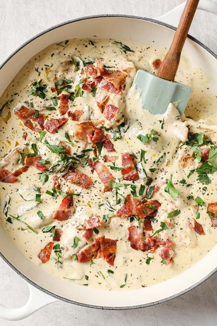 Overhead shot of a large pan with the One-Pan Creamy Dijon Chicken garnished with bacon crumbles, fresh parsley, and freshly-cracked pepper with a spatula for serving. The pan sits atop a marble countertop.