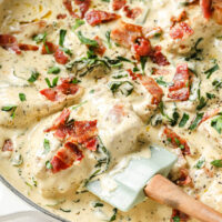 Overhead shot of a large pan with the One-Pan Creamy Dijon Chicken garnished with bacon crumbles, fresh parsley, and freshly-cracked pepper with a spatula scooping up a portion for serving. The pan sits atop a marble countertop.