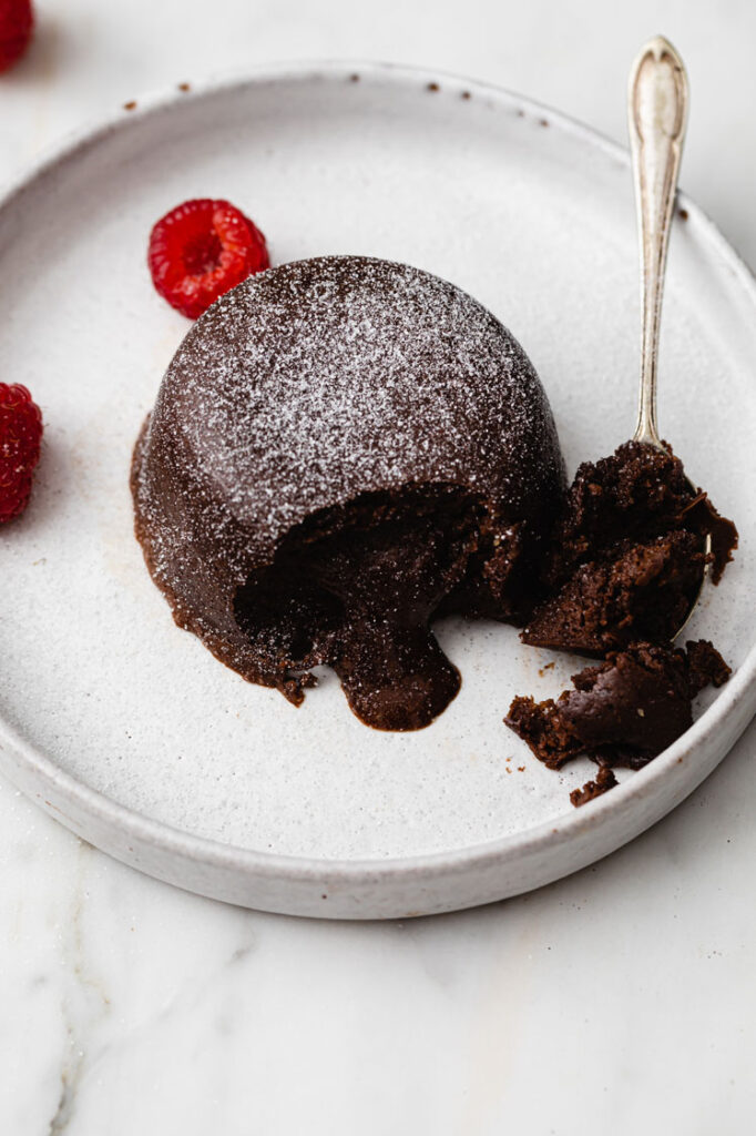 Overhead angled shot of the Instant Pot Lava Cake on a ceramic plate. A small portion is scooped up by a spoon, revealing the gooey inside of the lava cake. The lava cake is sprinkled with powdered monk fruit sweetener and garnished with a few fresh raspberries on the side. The plate rests atop a marble countertop.