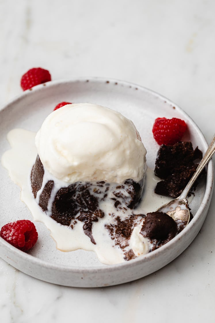 Overhead close-up shot of the Instant Pot Lava Cake on a ceramic plate. A small portion is scooped up by a spoon, revealing the gooey inside of the lava cake. The lava cake is sprinkled with powdered monk fruit sweetener and garnished with a few fresh raspberries on the side. The plate rests atop a marble countertop.