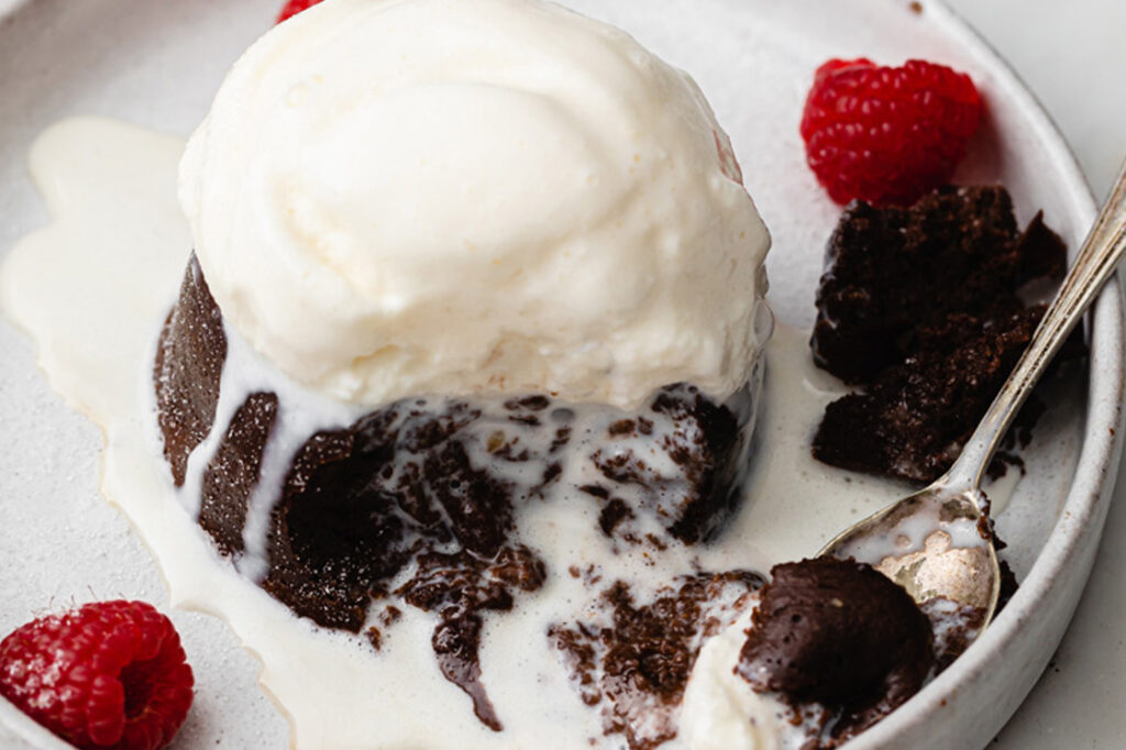 Close-up shot of the Instant Pot Lava Cake on a ceramic plate topped with keto-friendly ice cream, with the melted portion covering and spilling on the sides of the lava cake. A small portion is scooped up by a spoon, revealing the gooey inside of the lava cake, plus a few fresh raspberries on the side. The plate rests atop a marble countertop.