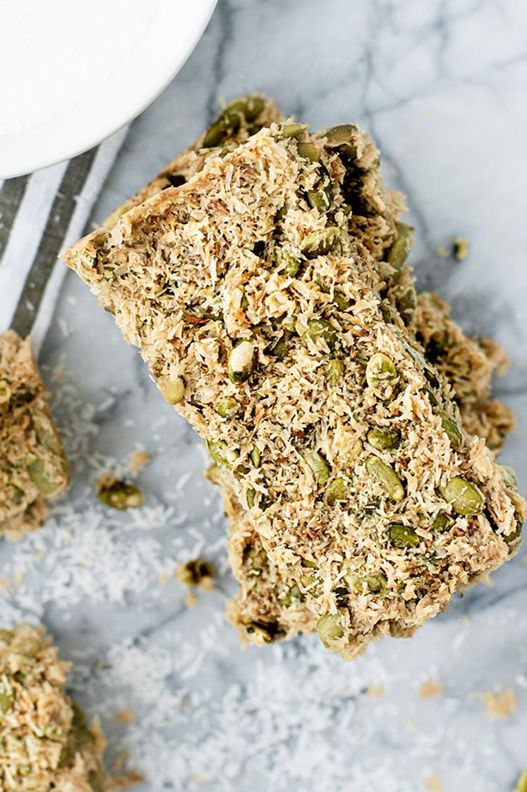 Overhead close-up shot of stacked Low Carb Granola Bars sprinkled with shredded coconut atop a marble countertop. There is also a bowl of monk fruit sweetener over a striped cloth table napkin in the background.