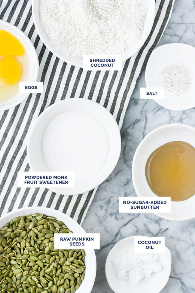 Overhead shot of labeled ingredients for the Low Carb Granola Bars in bowls over a striped cloth table napkin atop a marble countertop.