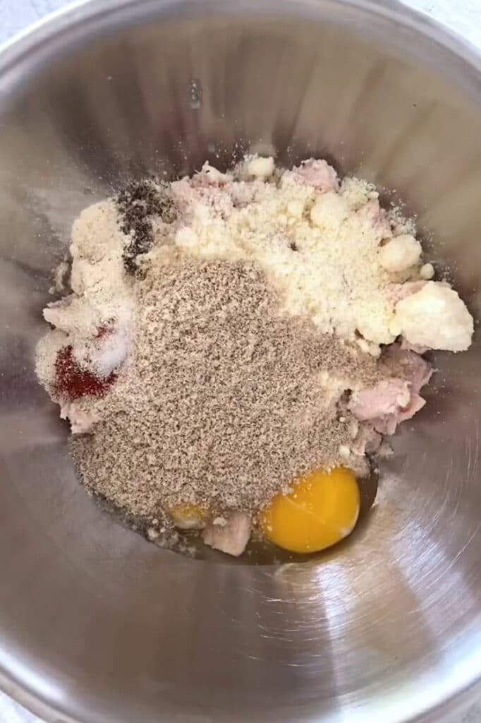 Overhead shot of canned chicken, eggs, grated Parmesan cheese, sunflower seed meal, garlic powder, onion powder, pepper, paprika, and salt in an aluminum mixing bowl.