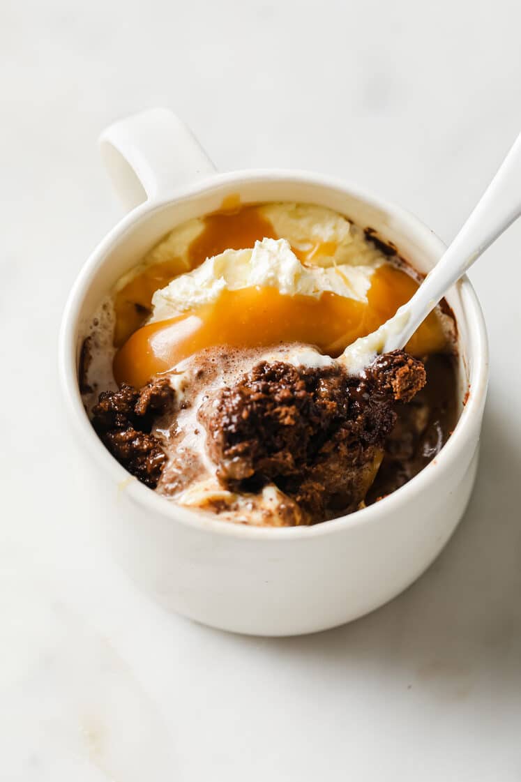 Overhead angled shot of a mug with the Low-Carb Double Chocolate Mug Cake garnished with a scoop of ice cream and drizzled with caramel. The mug has a spoon and rests atop a marble countertop.