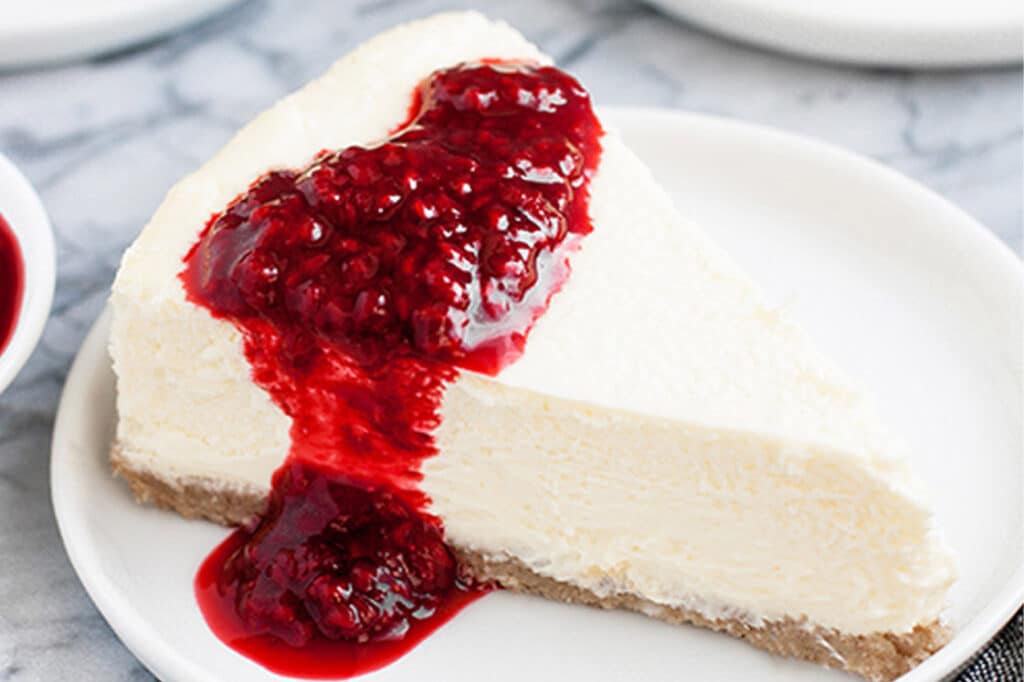 Overhead close-up shot of a slice of Nut-Free Keto Cheesecake drizzled with raspberry sauce served on a small plate atop a marble countertop.
