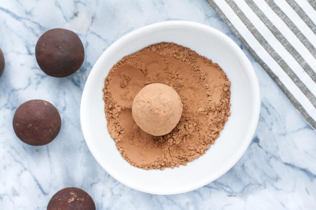 Overhead shot of one chilled Chocolate Truffle Fat Bomb on a shallow dish coated with cocoa powder and powdered monk fruit sweetener mixture, along with four chilled chocolate truffle fat bombs atop a marble countertop. In the background, you can also see a striped cloth table napkin.