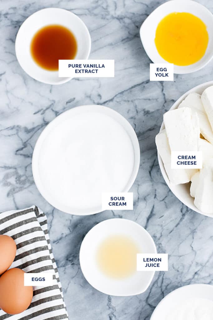Overhead shot of labeled ingredients for the cheesecake in small bowls atop a marble countertop.