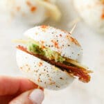 Close-up shot of one breakfast egg slider held over a marble plate with the remaining breakfast egg sliders. The breakfast egg sliders are skewered with a toothpick and garnished with paprika, freshly cracked pepper, and salt.