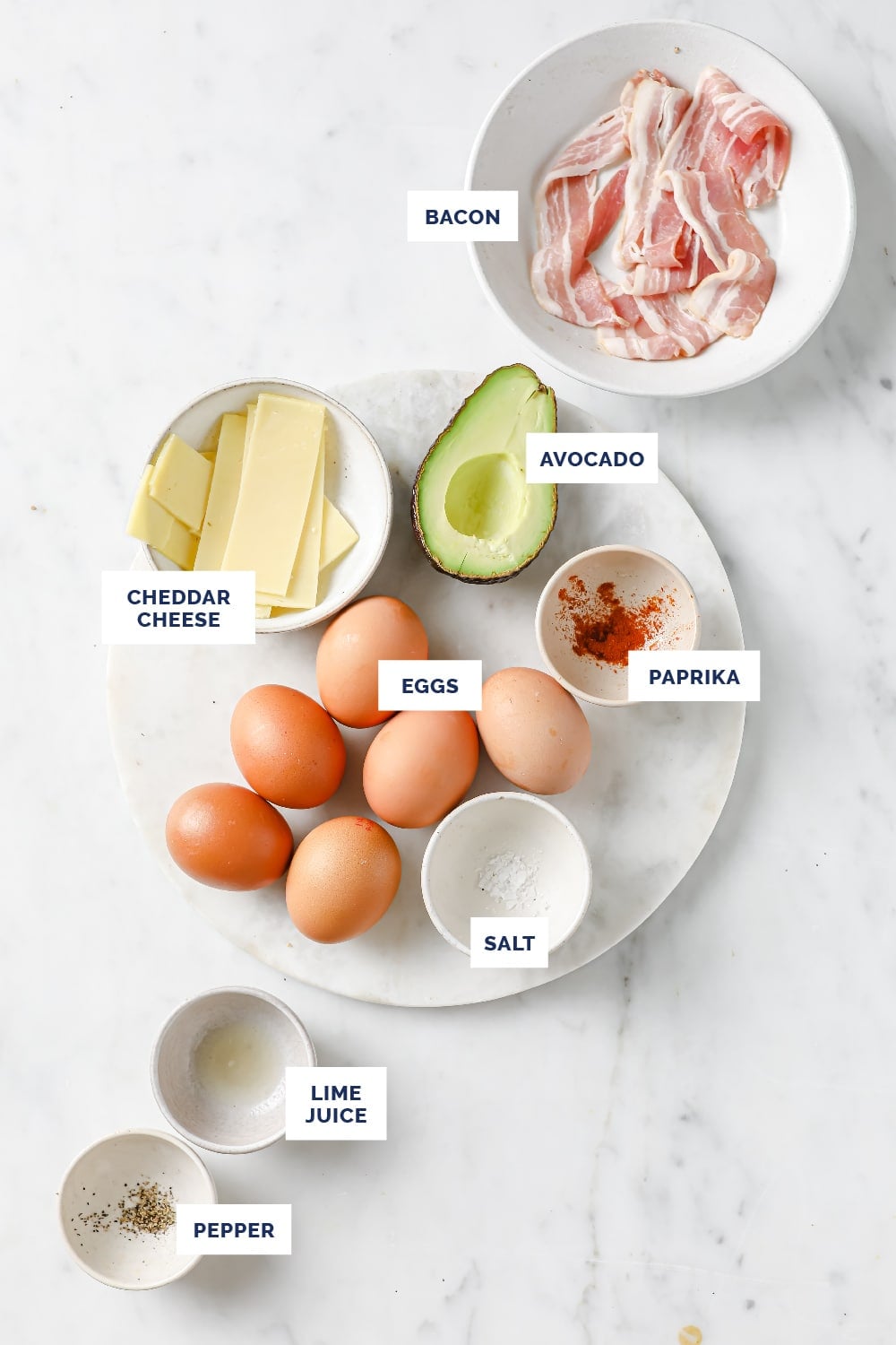 Overhead shot of labeled ingredients for the Breakfast Egg Sliders recipe in bowls and on a round marble cutting board atop a marble countertop.