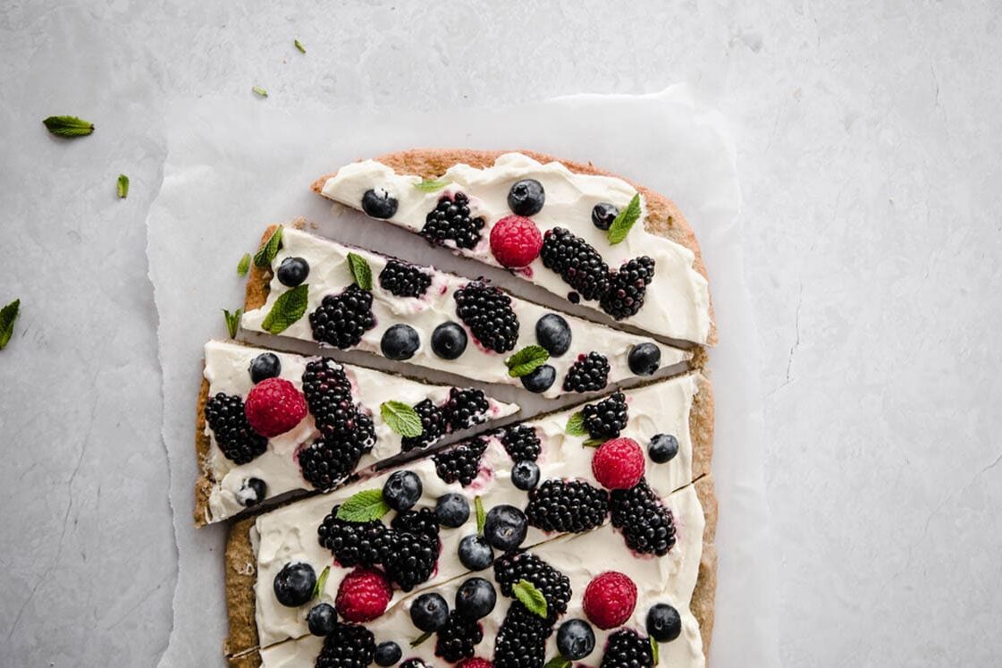 Fruit pizza atop parchment paper on a marble countertop sliced into pieces and ready to serve.