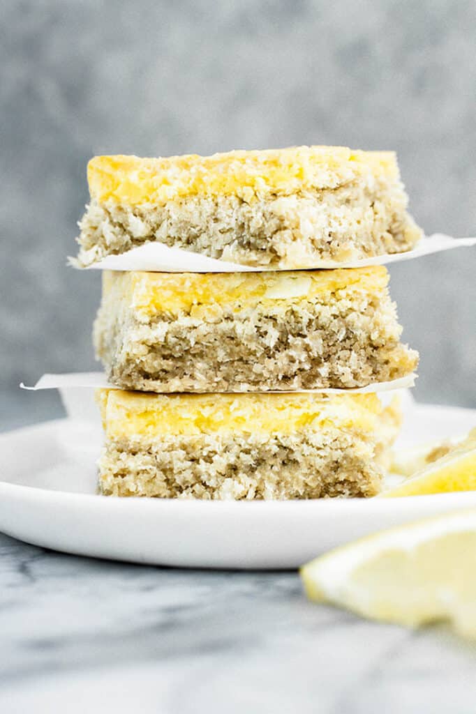 Three Keto Sugar-Free Gluten-Free Lemon Bars stacked and separated by parchment papers, with lemon slices on a ceramic plate.