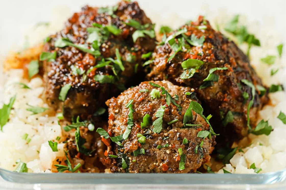 Close-up shot of steamed cauliflower rice and three meatballs drizzled with sauce and garnished with fresh parsley in a glass meal prep container atop a marble countertop.