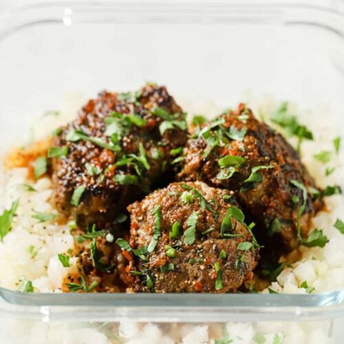 Steamed cauliflower rice and 3 meatballs with sauce and garnished with parsley in a glass meal prep container.
