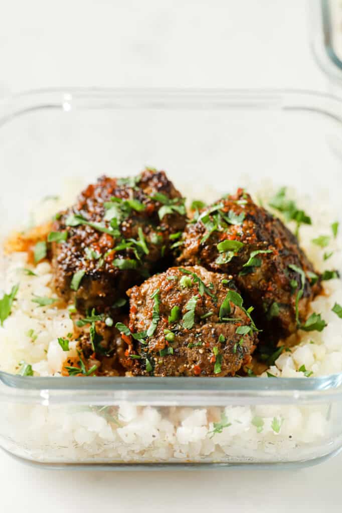Close-up shot of steamed cauliflower rice and three meatballs drizzled with sauce and garnished with fresh parsley in a glass meal prep container atop a marble countertop.