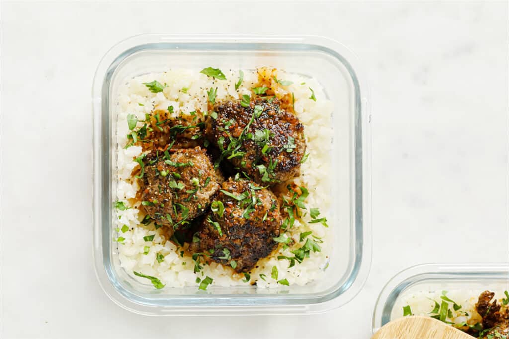 Overhead shot of steamed cauliflower rice and three meatballs, drizzled with sauce and garnished with fresh parsley in a glass meal prep container atop a marble countertop. In the background, there’s another meal prep container with cauliflower rice and meatballs.