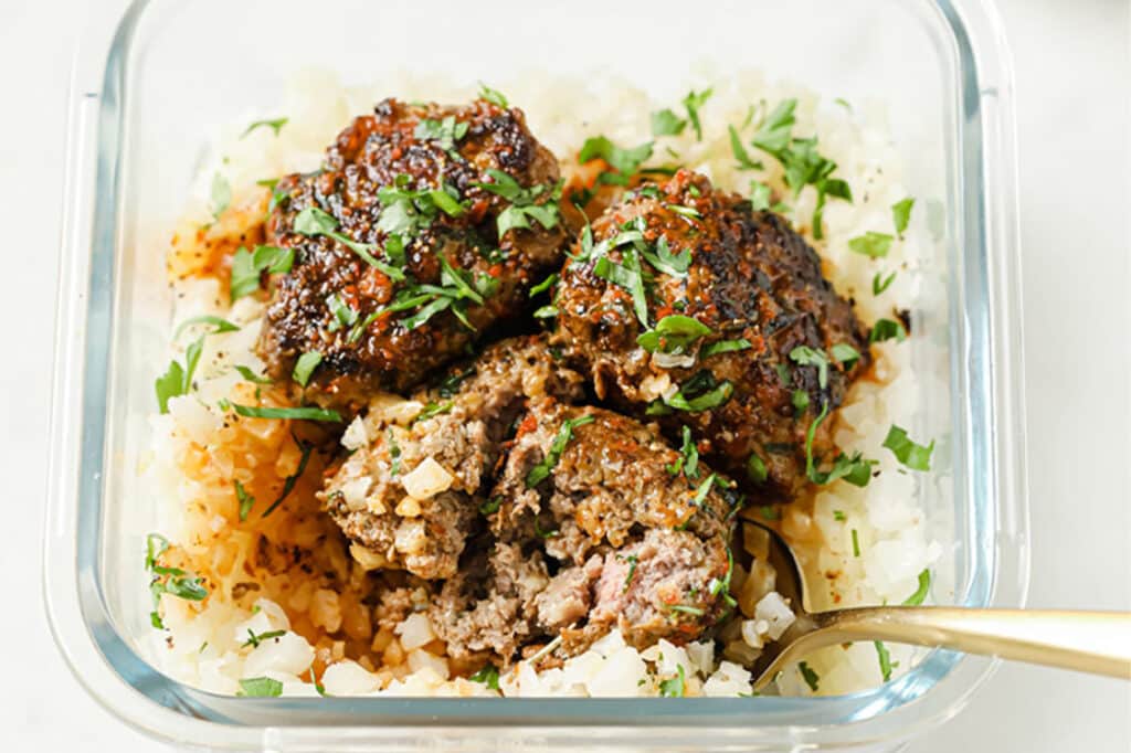 Overhead shot of steamed cauliflower rice and three meatballs, drizzled with sauce and garnished with fresh parsley in a glass meal prep container atop a marble countertop. One meatball is halved and spooned along with some cauliflower rice.