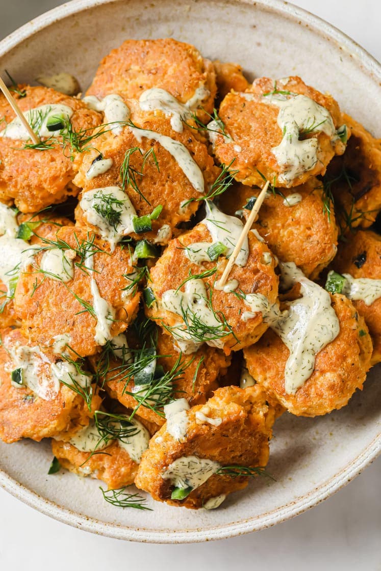 Overhead shot of a bowl with the spicy salmon poppers skewered with toothpicks, drizzled with aioli, and garnished with fresh dill and chopped jalapenos.