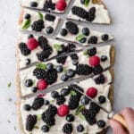 Hand holding knife slicing piece of fruit pizza on marble countertop.